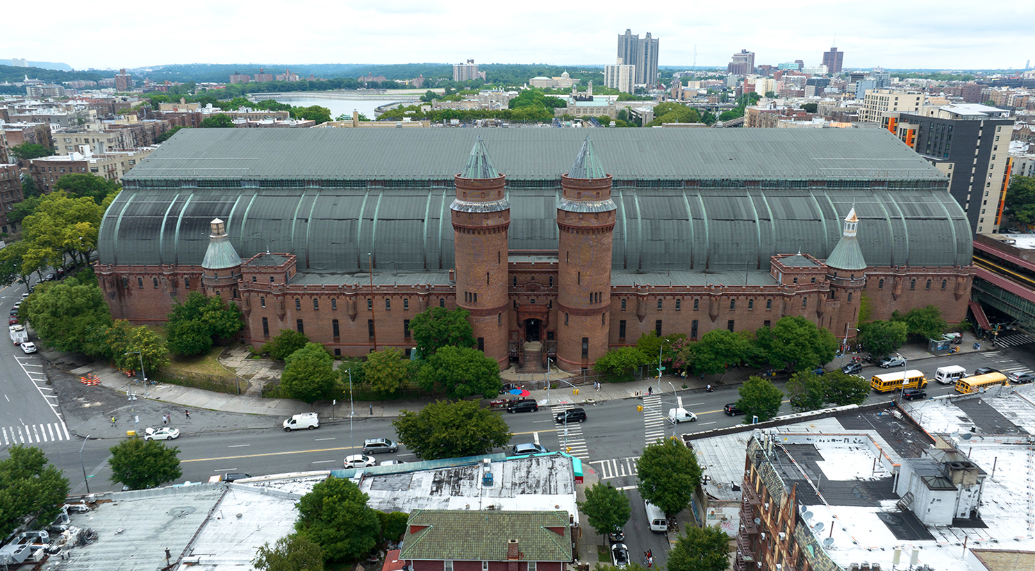 Aerial view of Kingsbridge Armory building with red brick walls and twin turrets, surrounded by NYC streets and greenery. The large, curved metal roof contrasts with the urban landscape, and several vehicles are visible on the roads nearby.