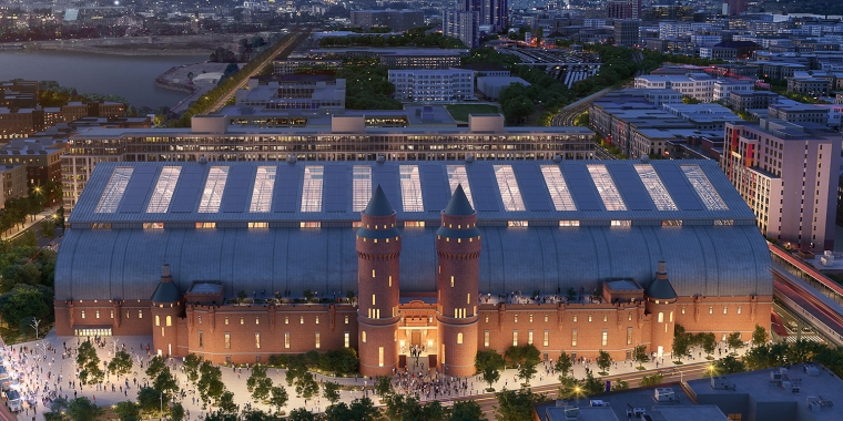 Aerial rendering of a large, illuminated Kingsbridge Armory building with two towers, surrounded by cityscape at dusk. The building features a glass-domed roof and is set against a cloudy sky with a pinkish hue. Streets and trees are visible around the structure. Image credit: FXCollaborative